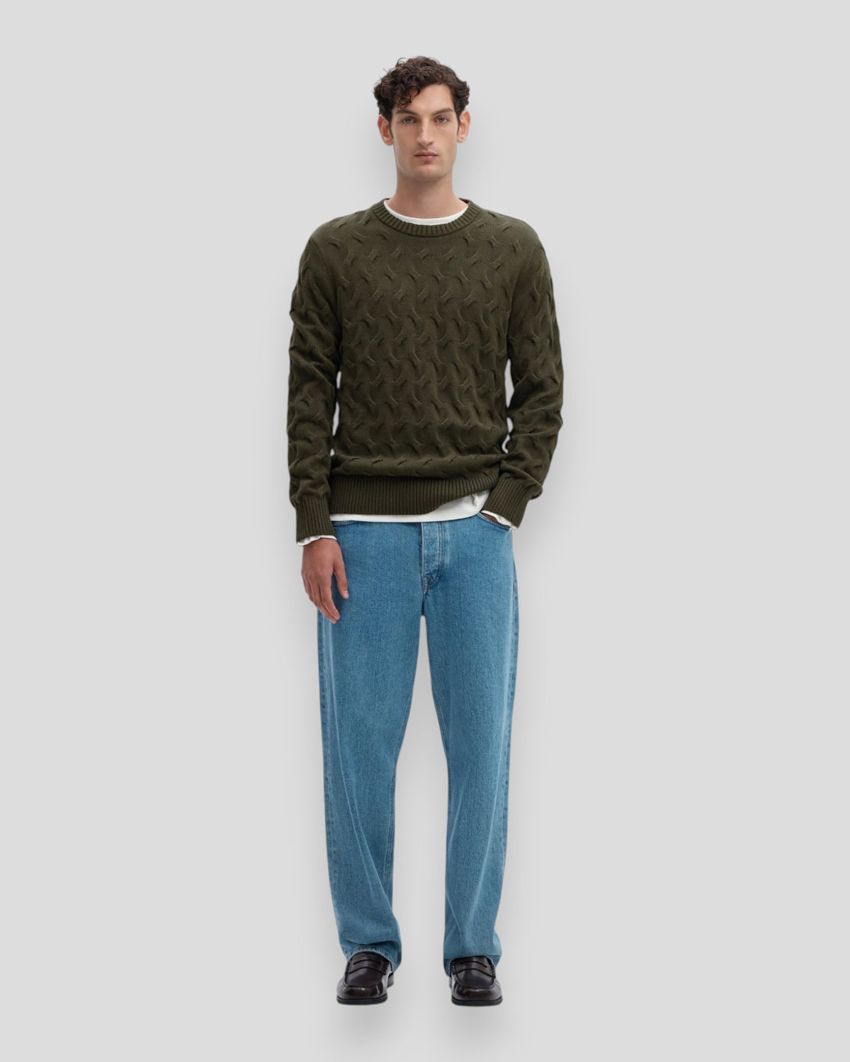 Mand i olivegrøn trøje med cableslyng og round neck, farve Forest Night, pari med blå jeans.